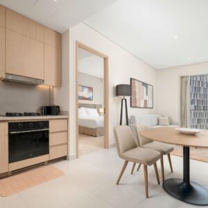 A bright and modern apartment interior featuring a clean kitchen with wooden cabinetry, a small dining table, and a view into a neatly made bedroom, showcasing high-end property standards.