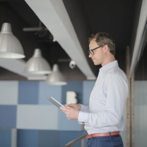 A professional man in a light blue shirt and glasses standing in a sunlit, modern office space while reviewing data on a digital tablet.