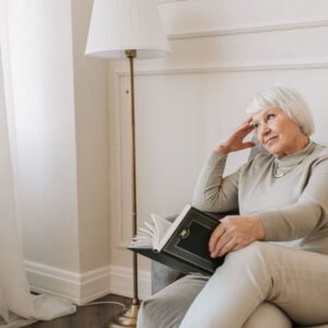 An older woman sitting in a modern, sunlit armchair, resting her head on her hand while holding a book, representing a peaceful and contemplative retirement lifestyle.