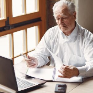 A senior man sitting at a desk with a laptop and smartphone, carefully reviewing and turning the pages of a notebook, representing financial planning or active management in retirement.