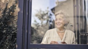 A candid and warm portrait of an elderly woman with a pearl necklace, smiling thoughtfully as she looks out of a window from inside a comfortable home.