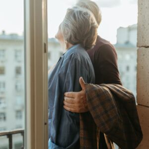 An older couple viewed from behind, standing together on a balcony and looking out over a city, with the man's arm around the woman, symbolizing a shared future and "rightsizing" into urban living.