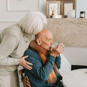 A touching scene of an elderly woman leaning down to kiss the head of an elderly man who is sitting and drinking from a clear glass, set in a bright, warm living room.