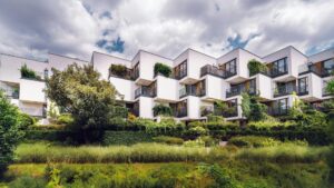 A wide-angle view of a modern, white multi-unit residential building with unique staggered balconies, surrounded by dense green ivy and professional landscaping under a cloudy sky.
