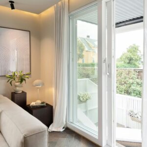 An interior view of a bright, minimalist living space looking through open white sliding glass doors toward a sunlit balcony and a neighboring yellow house.