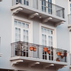 A vertical architectural shot of a white classic-style building facade featuring multiple balconies with black wrought-iron railings and several terracotta potted plants.