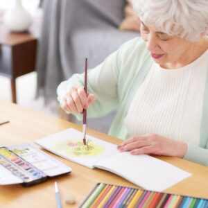 an elderly woman with short white hair peacefully painting a watercolor of a flower in a notebook on a wooden table.