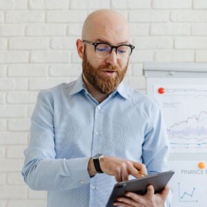 A professional, bearded man wearing glasses and a light blue dress shirt, focused on a digital tablet while standing in front of a whiteboard displaying financial growth charts.